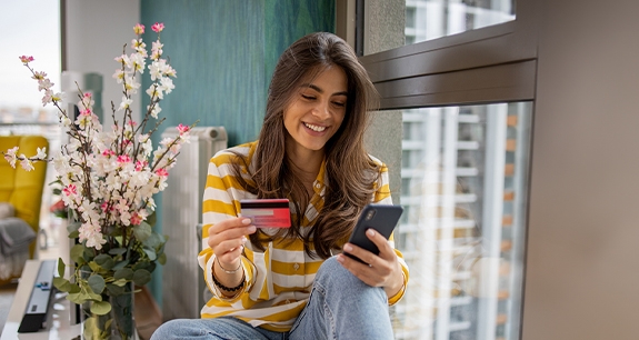 ragazza sorridente che guarda il cellulare con anche la carta di credito seduta sul davanzale della finestra | Crédit Agricole ragazza sorridente che guarda il cellulare con anche la carta di credito seduta sul davanzale della finestra | Crédit Agricole
