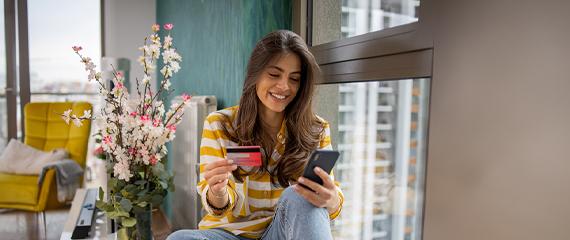 ragazza sorridente che guarda il cellulare con anche la carta di credito seduta sul davanzale della finestra | Crédit Agricole ragazza sorridente che guarda il cellulare con anche la carta di credito seduta sul davanzale della finestra | Crédit Agricole