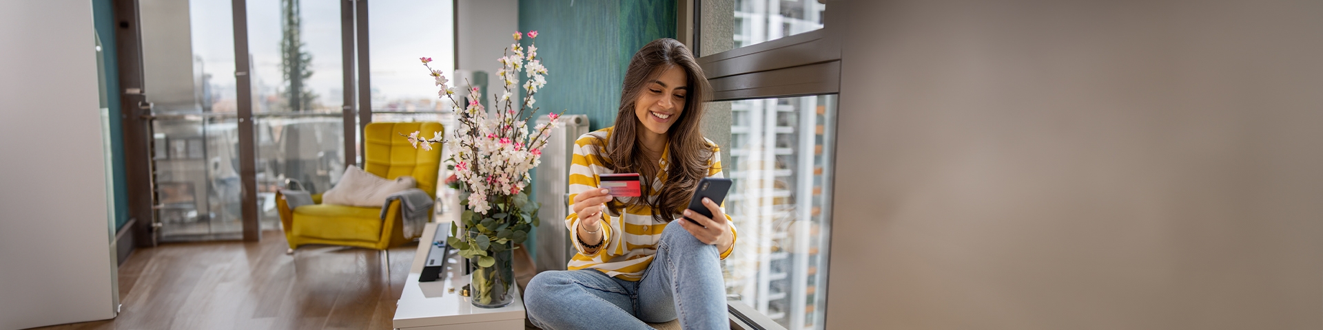 ragazza sorridente che guarda il cellulare con anche la carta di credito seduta sul davanzale della finestra | Crédit Agricole ragazza sorridente che guarda il cellulare con anche la carta di credito seduta sul davanzale della finestra | Crédit Agricole