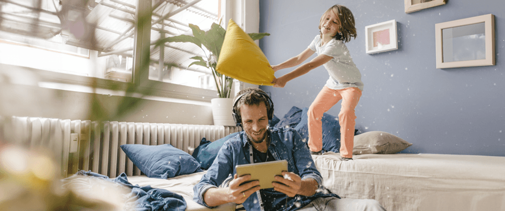 papà che guarda il tablet con le cuffie e bambina dietro di lui sul divano che sta per colpirlo con un cuscino | Crédit Agricole papà che guarda il tablet con le cuffie e bambina dietro di lui sul divano che sta per colpirlo con un cuscino | Crédit Agricole