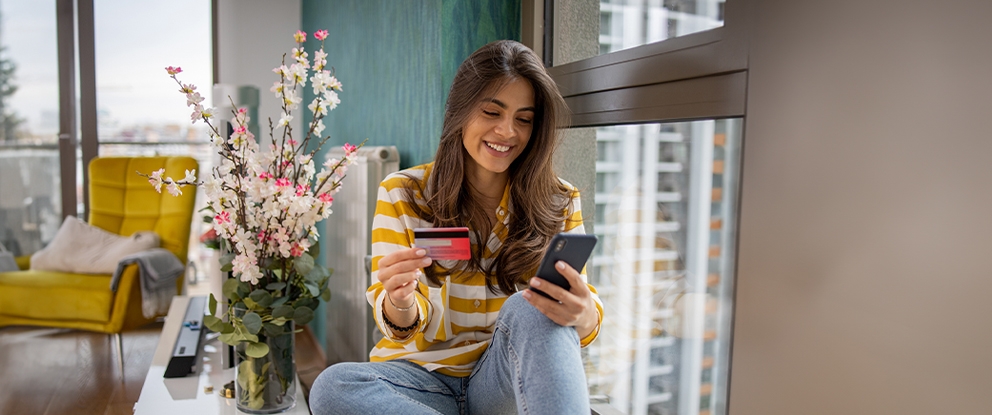 ragazza sorridente che guarda il cellulare con anche la carta di credito seduta sul davanzale della finestra | Crédit Agricole ragazza sorridente che guarda il cellulare con anche la carta di credito seduta sul davanzale della finestra | Crédit Agricole