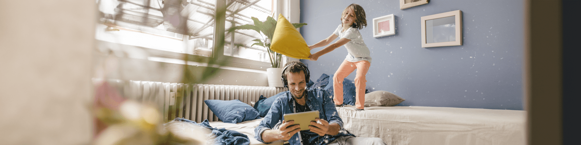 papà che guarda il tablet con le cuffie e bambina dietro di lui sul divano che sta per colpirlo con un cuscino | Crédit Agricole papà che guarda il tablet con le cuffie e bambina dietro di lui sul divano che sta per colpirlo con un cuscino | Crédit Agricole