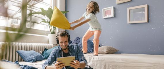 papà che guarda il tablet con le cuffie e bambina dietro di lui sul divano che sta per colpirlo con un cuscino | Crédit Agricole papà che guarda il tablet con le cuffie e bambina dietro di lui sul divano che sta per colpirlo con un cuscino | Crédit Agricole
